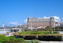 Biarritz | L’avenir incertain de l’Hôtel du Palais : la vente en question ? Famous hotel of Biarritz against azure sky. There is square with green park in the foreground. There are cliff with lighthouse and ocean waves surging in the background.