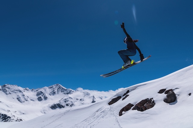 Young female skier jumping from ski jump on sunny day in snowy alpine mountains, moment in flight.
