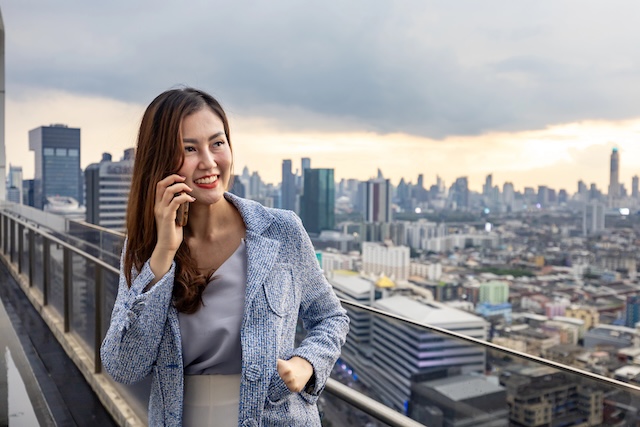 Asian business woman in formal suit is phone calling customer while standing outside skyscraper building for marketing, connection, corporate work, real estate, housing and urban development