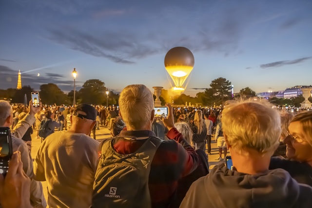 Paris, France - 07 28 2025: People standing from behind take photos with cellphones of the Olympic Cauldron as it takes flight from the Tuileries Garden at sunset.